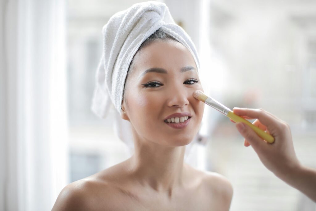 Smiling woman with a towel on her head enjoying a makeup session indoors, highlighting natural beauty and healthy skin.