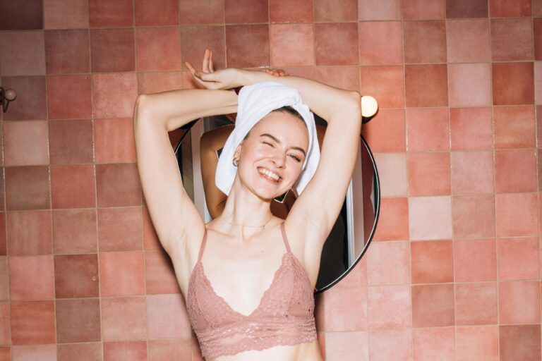 A cheerful woman with a towel on her head posing in a pink tiled bathroom, smiling blissfully.